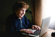 An older woman sits at a desk with a laptop, her finger poised over the keyboard as she works.