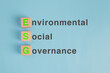 A wooden block on pastel blue paper background with the green letters ESG written on it. Concept of Environmental, social and governance principle for public responsibility.