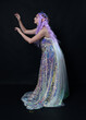 Full length portrait of beautiful female model with long purple hair wearing elf ears, a fantasy fairy crown and  rainbow glitter sequin ball gown.  walking away, back view, isolated dark studio 