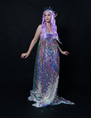 Full length portrait of beautiful female model with long purple hair wearing elf ears, a fantasy fairy crown and  rainbow glitter sequin ball gown. graceful standing pose,  isolated in dark studio bac