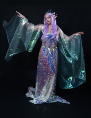 Full length portrait of beautiful female model with long purple hair wearing elf ears, a fantasy fairy crown and  rainbow glitter sequin ball gown. graceful standing pose,  isolated in dark studio bac