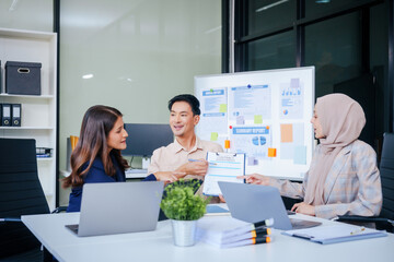 A diverse group of professionals, including a male Asian businessman and a female Muslim businesswoman in hijab, gather around a desk equipped with tablets and laptops for a productive meeting.