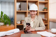© Ekkasit A Siam - Asian female architect wearing white hard hat, looking at smartphone while sitting at desk in modern office. Professional workspace with wooden shelves, green plants, and documents visible.