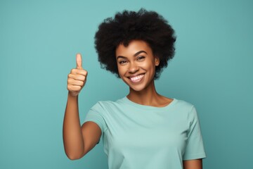 Wall Mural - Portrait of a smiling afro-american woman in her 30s showing a thumb up in front of pastel teal background
