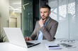 © Liubomir - Young businessman working on a laptop in a modern office. He appears to be thinking and planning, with notes and devices on the desk.