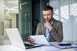 © Liubomir - Businessman deeply focused on analyzing documents at office desk with laptop and pen, showcasing office work and determination.