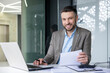 © Liubomir - Smiling businessman at his desk working on laptop and checking documents in a contemporary office setting.