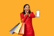 © Prostock-studio - A joyous young woman dressed in a vibrant red dress stands against an orange background, holding colorful shopping bags in one hand and presenting a blank-screen smartphone mockup