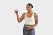 © Prostock-studio - A happy young black woman stands confidently with a tape measure around her waist, holding a clear glass of water. Her activewear suggests a focus on health and fitness