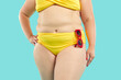 Crop shot of overweight, obese woman with fat belly wearing yellow bikini bottom and bra with red sunglasses standing with hand on hip isolated on blue background. Beach swimwear, summer concept