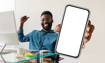 Poster - African American man sitting at workdesk with pc, holding a cell phone in his hand, showing blank white screen and gesturing