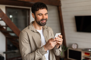 Aufkleber - A man is standing in a living room, holding a cell phone in his hand and looking at the screen intently. The room is well-lit, with furniture and decor visible in the background.