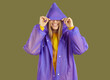 Smiling young woman with hood on her head hiding from rain. Portrait of happy young blonde woman dressed lilac raincoat standing over isolated studio background