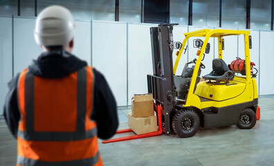 Poster - Forklift behind man. Guy warehouse employee with back to camera. Forklift for unloading work. Logistician in orange vest and helmet. Forklift with cardboard boxes in industrial building. Art focus