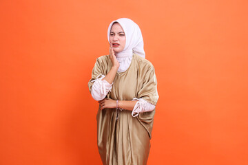 expression of a beautiful indonesia girl, candid, sad, aching teeth, holding her cheek, wearing a kaftan Muslim dress with a hijab, isolated on an orange background. Concept of health care, promotion