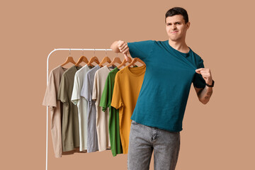 Wall Mural - Handsome young displeased man showing thumb-down gesture and rack with different t-shirts on brown background