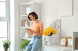 © Pixel-Shot - Young woman using mobile phone with daffodils in vase on commode at home