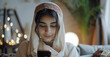 © Artyom - Young Middle Eastern woman in hijab using smartphone at home, surrounded by cozy decor
