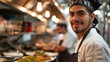 © khonkangrua - A confident male chef smiling at the camera while preparing food in a busy commercial kitchen with his team.