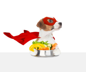 Aufkleber - Funny Jack russell terrier puppy wearing superhero costume holds bowl of vegetables above empty white banner. Isolated on white background