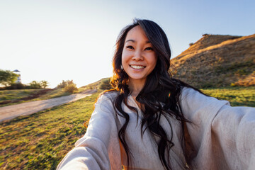 Wall Mural - A young attractive Asian woman in a linen dress takes a selfie and laughs outdoors