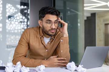 Stressed businessman working at desk with crumpled papers and laptop in office