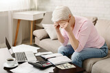 Fototapeta  - A senior woman is seated on a couch with her hands covering her face, showing signs of stress or headache, surrounded by financial documents, a calculator, money, and a laptop computer