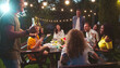 © ihorvsn - Joyful mixed-race friends having fun at garden party in evening. Caucasian guys and girls drinking and chatting outdoor. African American girl smiling. Asian female laughing. Summertime concept