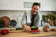 © Jelena - A happy adult man making a fresh salad and taking a cherry tomato while leaning on the kitchen island