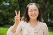 Happy blind middle aged asian woman pointing 2 fingers up, concept of v for victory, concept image for blind people, visual impair, accessibility, visual A11Y in summer green urban park