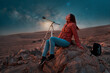© oscargutzo - beautiful woman sitting in the desert alone next to a telescope at night watching the starry sky, astronomy and stargazing concept