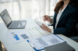 © Wasana - A woman in a business suit is sitting at a desk with a laptop and a calculator. She is looking at a piece of paper with numbers on it