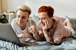 © Bliss - Two people, a lesbian couple with short hair, relax and connect on a bed while looking at a laptop screen.