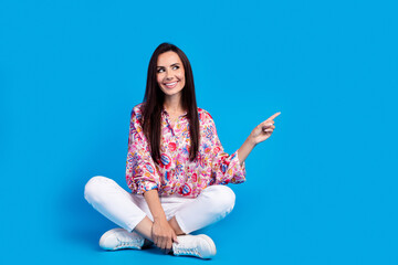 Poster - Full length photo of cute dreamy lady dressed print shirt sitting legs crossed pointing empty space isolated blue color background