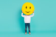 Full body photo of cheerful kid hiding face with handmade smile emoji isolated cyan color background