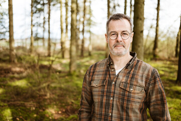 portrait of a middle-aged man standing in a serene forest wearing a plaid shirt and glasses, exuding