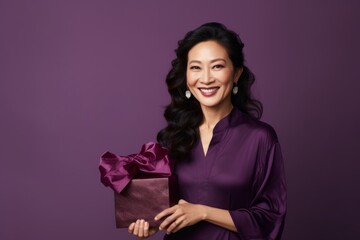Sticker - Portrait of a merry asian woman in her 40s holding a gift isolated on soft purple background