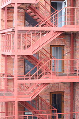 Poster - A red fire escape staircase next to a brick building. Suitable for urban architecture concepts