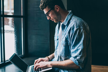 Caucasian male freelancer sitting near cafeteria window and typing new article in modern laptop application, young man blogger browsing wireless internet and updating profile in social networks