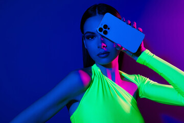 Portrait of gorgeous confident model girl hand hold smart phone cover one eye isolated on blue neon background