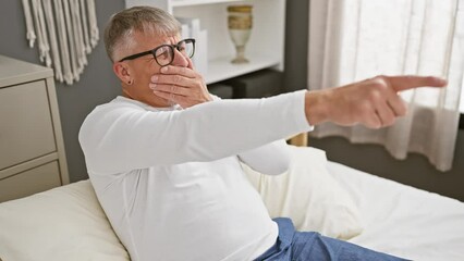 Wall Mural - Cheerful middle-aged grey-haired man playfully pointing a finger at you, wearing pyjamas, laughing with hand over mouth in a cosy bedroom