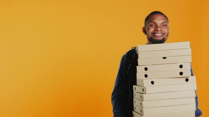 Wall Mural - African american pizzeria deliveryman holding stack of pizza boxes to deliver food order to customer in studio. Friendly takeout service employee carries takeaway meal package. Camera B.