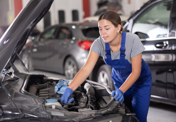 Wall Mural - Young female mechanic in uniform repairs under hood of car in car service station