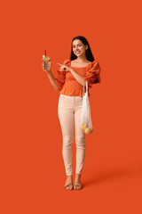 Poster - Beautiful young happy woman holding glass of fresh lemonade and string bag with citruses on orange background