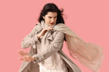 Wall Mural - Young woman in trench coat with scarf and strong wind on pink background