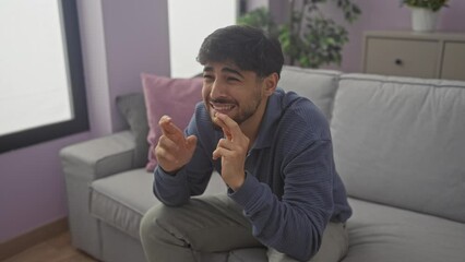 Sticker - Young man with beard sitting on couch at home crossing fingers for good luck and celebrating excitingly with a joyful expression.