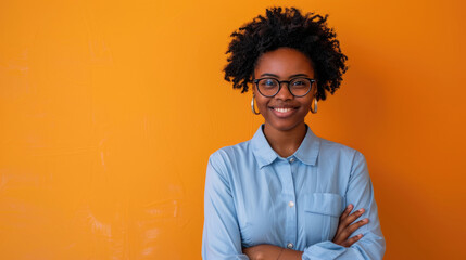 Happy Afro businesswoman in light blue shirt and stylish glasses standing with crossed arms on orange background, smiling friendly at the camera