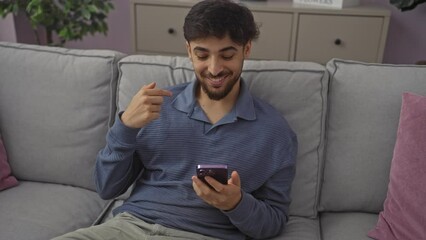 Wall Mural - Young, arab man, sitting at home on a sofa, happily pointing at self with a proud smile while using a smartphone