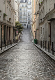 Fototapeta Uliczki - Perspective view of one of the old montmartre streets in Paris. A narrow cobblestone street with typical French architecture descends and rises, The historic district of Montmartre in the city of Pari