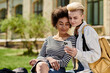 © Bliss - lesbian couple in casual outfits sit on the grass, engrossed in phone, chatting and sharing moments.